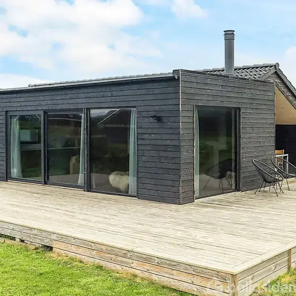 Sort træhus med store glaspartier står på en bred træterrasse. To stole og et bord står på terrassen. Omgivet af græsplæne og en klar himmel i baggrunden.