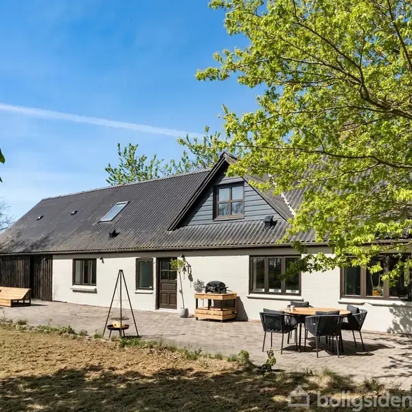 Et hvidt hus med sort tag ligger i en grøn have. Der er havemøbler og et hængegyngestativ på en flisebelagt terrasse omgivet af træer og græs.