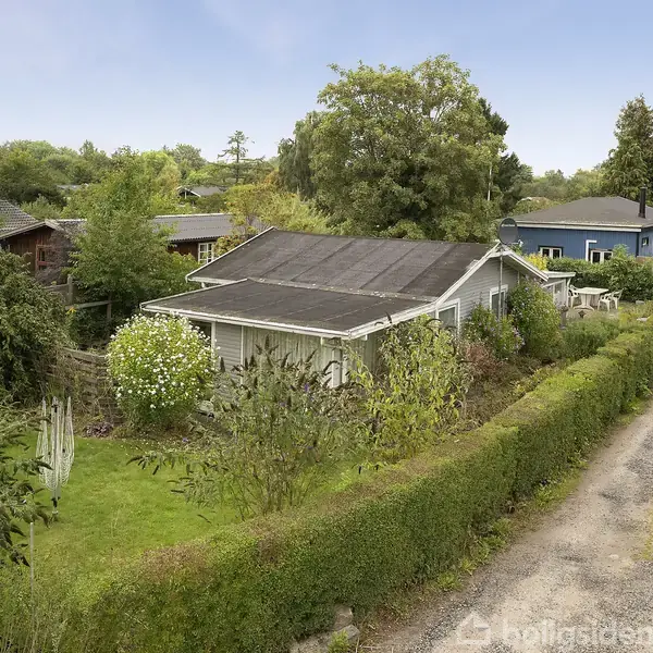 Et lille hus med have omgivet af tæt bevoksning og hække, beliggende langs en grusvej i et grønt og frodigt område med andre lignende huse i baggrunden.