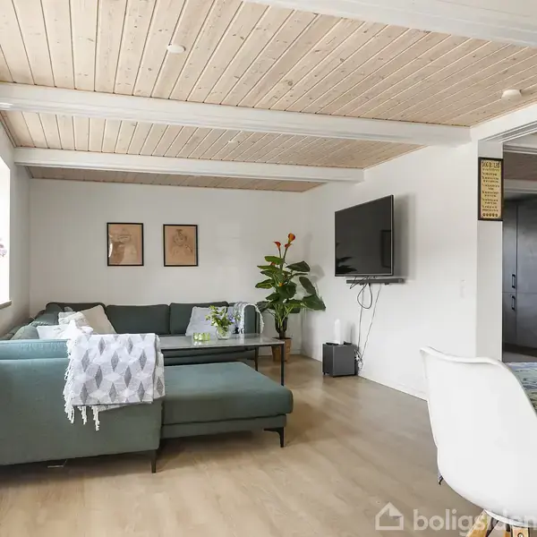En moderne stue med grøn L-formet sofa ved siden af et sofabord. Et tv hænger på væggen foran sofaen. Rummet har trægulv, lofter og lyse vægge.