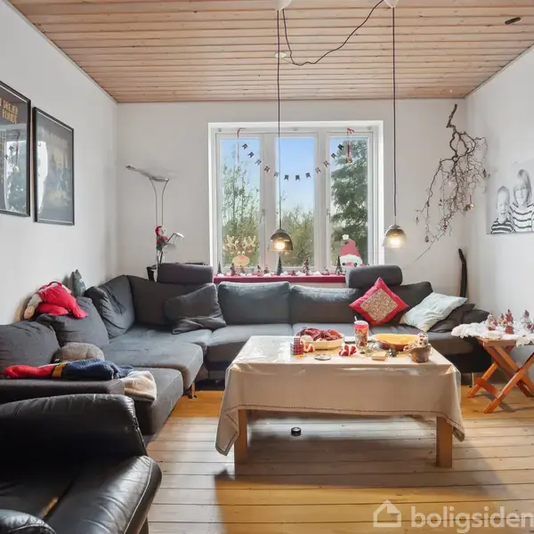 Stue med mørkegråt hjørnesofa, dækket bord og lænestol. Vindue med udsigt til have baggrund. Julepynt på bord og vindueskarm. Træloft og trægulve i en hyggelig atmosfære.