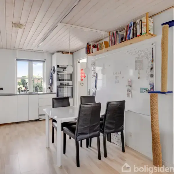 Køkken med hvidt bord og fire sorte stole; over bordet en hylde med bøger. Et vindue til venstre og hvide skabe langs væggene, med lys trælaminatgulv og loft.