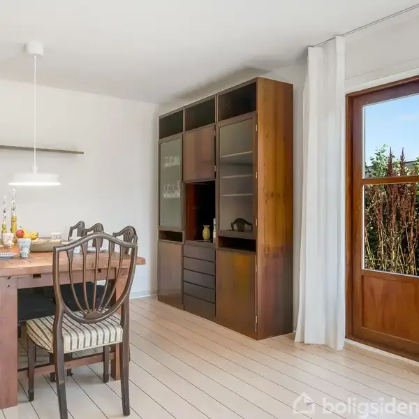 Spisebord med seks stole står i et lyst rum ved siden af en stor reol. Gennemsigtige døre i træ fører ud til en have. Bordet er pyntet med frugt og blomster.