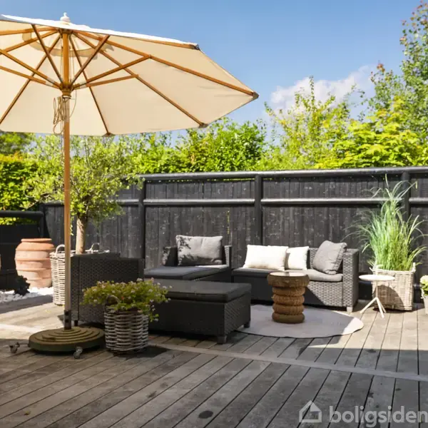 En terrasse med træplanker, havemøbler og en stor parasol. Møblerne inkluderer en sofa og puder. Baggrunden har sorte hegn og grønne planter. Himmel er blå med skyer.