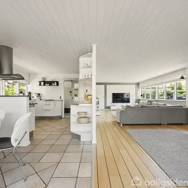 Et åbent stue-køkken område med moderne hvide møbler. Et rundt spisebord står nær køkkenet med emhætte. I stuen står en grå sofa foran et stort vindue og en TV-skærm.