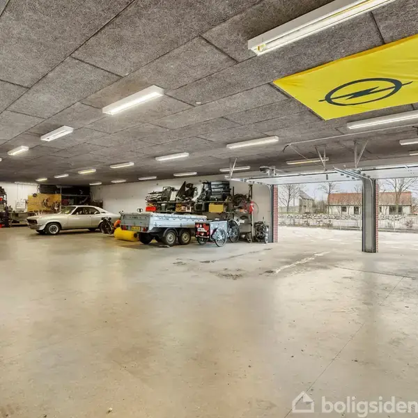 En garage med flere biler, inklusive en klassisk bil og en trailer med udstyr. Der er betongulv, loftslamper, og en gul Opel-flag hænger fra loftet. Udenfor ses en indkørsel.