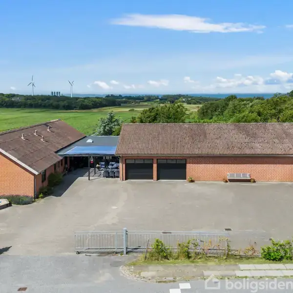 Rødt murstenshus med et fladt tag og garageporte. En campingvogn er parkeret foran. Omgivet af en stor, åben gårdsplads med en grøn mark og vindmøller i baggrunden.