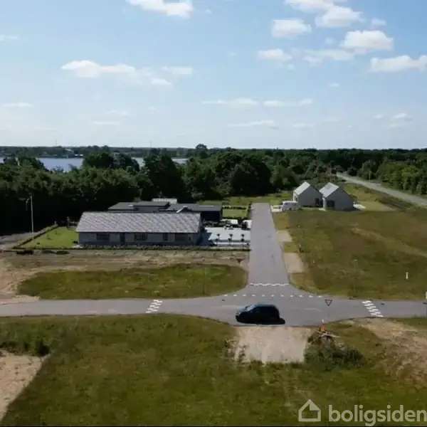 Bygningerne står langs en asfaltvej omgivet af grønne arealer og skov, mens en bil holder ved et kryds i forgrunden. En sø er synlig i baggrunden.