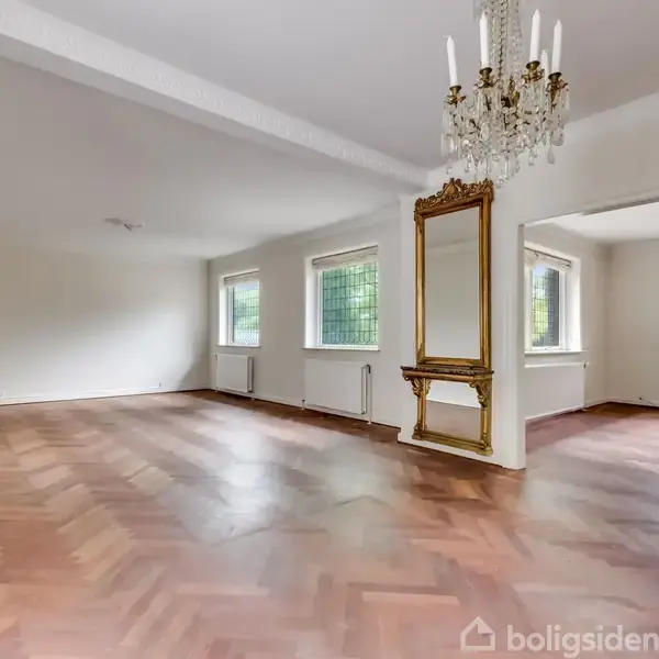 Stort, tomt rum med parketgulv og to elegante lysekroner, skilt af en væg med et stort, guldbelagt spejl. Flere vinduer giver naturligt lys.