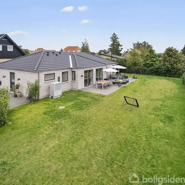 Et moderne hus med fladt tag står stille ved siden af en stor, grøn græsplæne. Haven er omgivet af buske, og terrassen har et bord med parasol.