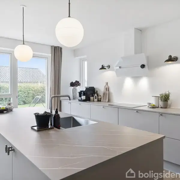 Køkken med ø, hvor der står en sort skål, omgivet af moderne apparater og hvide skabe. Store vinduer med udsigt til en have giver rummet lys.