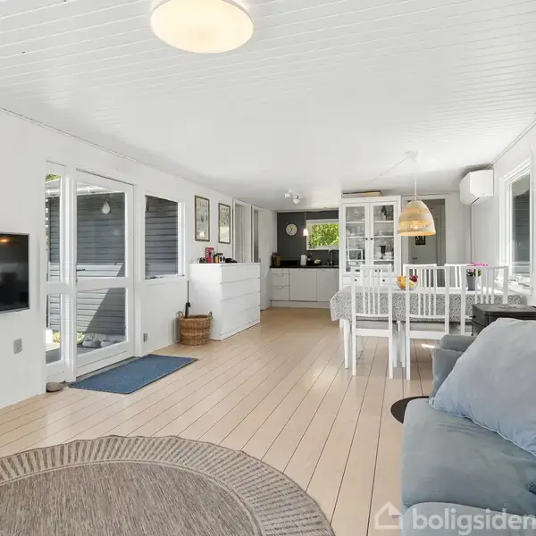Stue med sofa til højre, fladskærms-tv på væggen, rundt tæppe på gulvet. Spisebord med stole til venstre, køkken i baggrunden. Lyst interiør med vinduer og lyse trægulve.