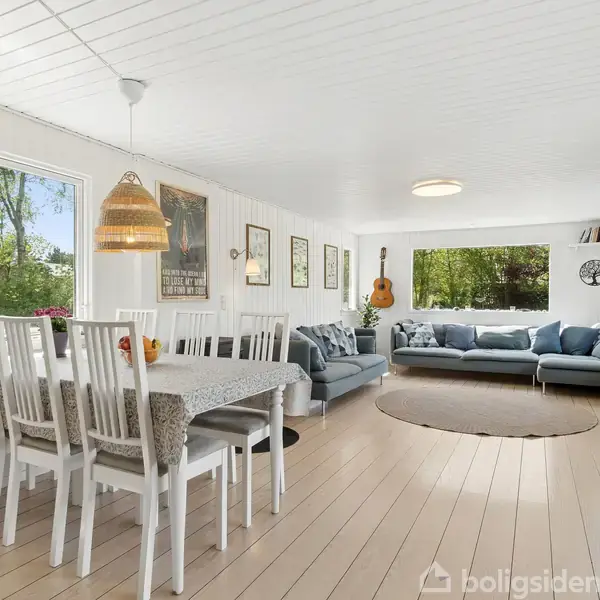 Spisestuemøbler står langs et træbord med hvide stole. I stuen er en stor grå sofa placeret foran et stort vindue. Væggene har kunstværker, og en guitar hænger til dekoration.
