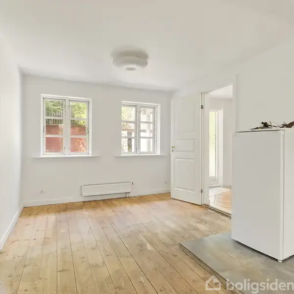 Et tomt rum med trægulv og hvide vægge, rummet har to vinduer og en dør. En hvid radiator under vinduerne og en lille hvid reol står ved døren.