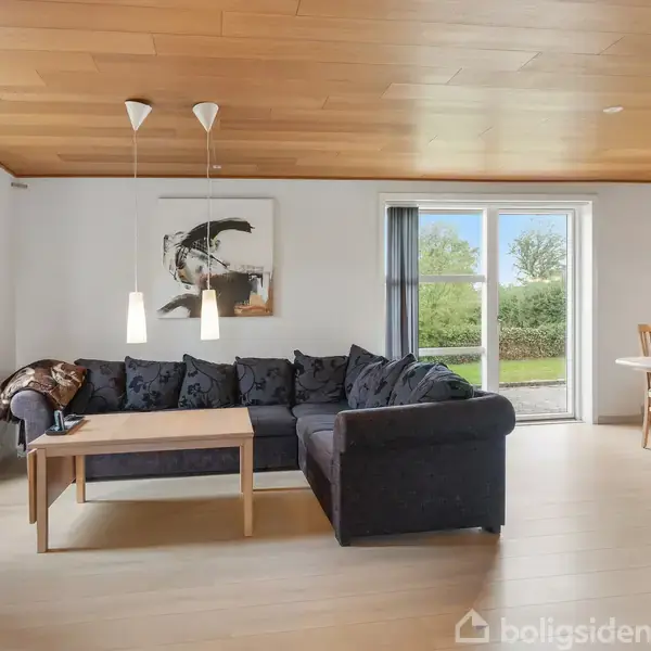 Sort hjørnesofa står i en stue med trægulv. To lamper hænger over et træbord foran sofaen. En stor glasdør fører til en grøn have. En spisebord med stole står til højre.