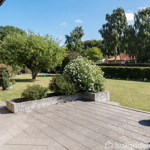 Terrasse med flisebelægning omgivet af velplejede buske og grøn græsplæne. Bevoksningen inkluderer store træer i baggrunden. Klart solskinsvejr skaber en lys og indbydende atmosfære.