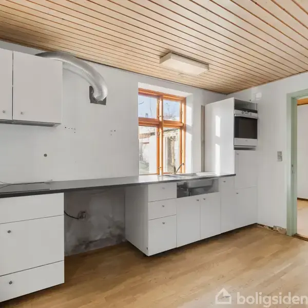 Køkken med hvide skabe og sorte bordplader. Et vindue giver lys ind, og der er en ovn indbygget. Loftet har træpaneler, og gulvet er lyst træ.