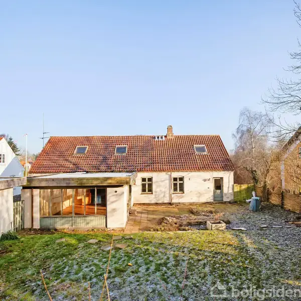 Et hvidt hus med rødt tegltag står i en have med græsplæne og træer. Der er flere vinduer på taget, og omgivelserne er fredelige og grønne.