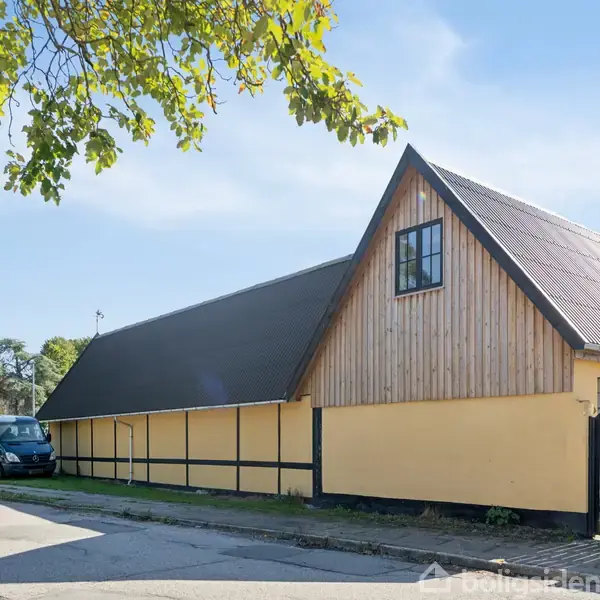 En gul bygning med sort tag står ved en vej, omkranset af træer. I baggrunden ses et kirketårn. En bil er parkeret til venstre af bygningen.