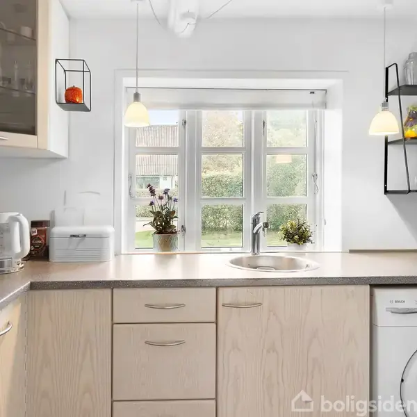 Et køkken med lyse træskabe, en vask, og en vaskemaskine. Hylder med plantede dekorationer hænger ved et vindue, der lukker naturligt lys ind. En elkedel er placeret på bordpladen.