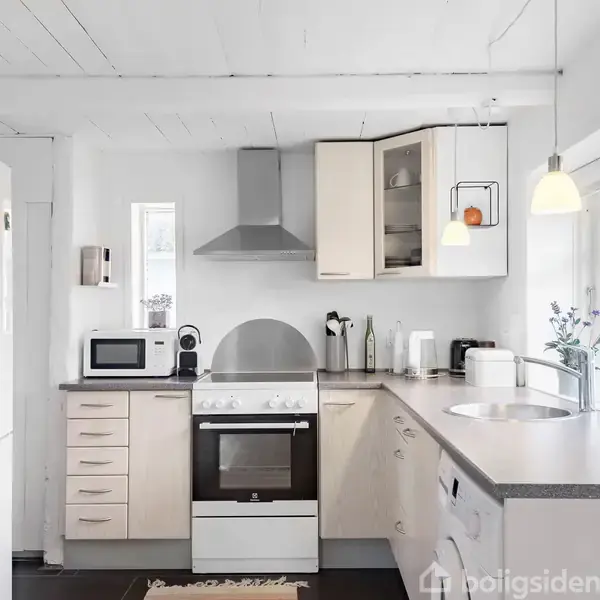 Et moderne køkken indeholder et komfur med emhætte, omgivet af hvide skabe og grå bordplader. En mikroovn og hvide køleskab står ved siden af. Lyse, hængende lamper lyser rummet op.