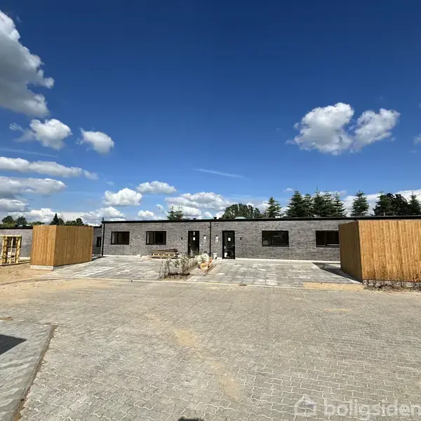 Rækkehuse står ubenyttede på en nyligt belagt stenterrasse. Himlen er blå med spredte skyer, og buske ses i baggrunden.