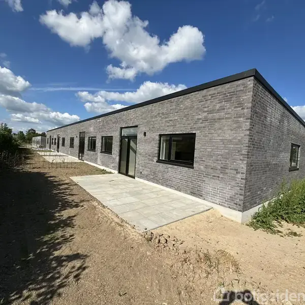 En lang, lav bygning af grå mursten med flere vinduer og et hovedindgang. Bygningen står på en flisebelagt plads omgivet af jord og grønt landskab under en himmel med hvide skyer.
