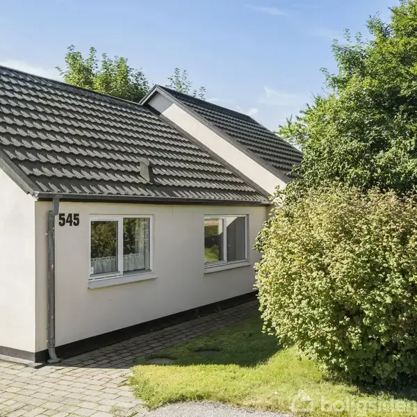 Et hvidt hus med sort tag står stille i sollys omgivet af grønne træer og buske. På husets facade står tallet "545". Huset ligger på en flisebelagt grund.