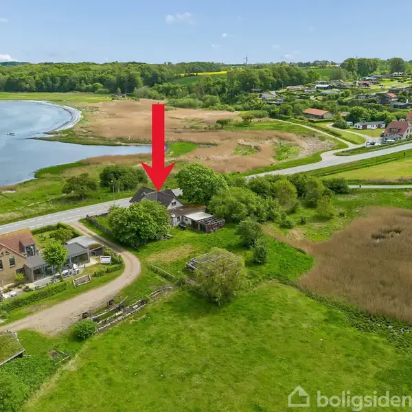 Hus omgivet af grønt område med en rød pil, der peger på det; beliggende nær en sø med en sti og omkringliggende huse i baggrunden.
