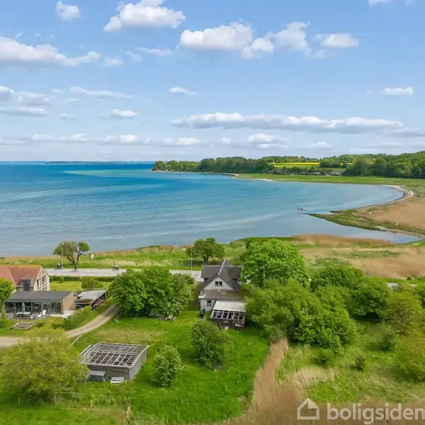 Huse ligger ved en kyststrækning med grønne områder og stranden. De omgive træer og marker, mens havet strækker sig under en blå himmel med hvide skyer.