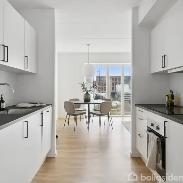 Et moderne køkken med hvide skabe og sorte greb strækker sig mod en spisestue i baggrunden. Spisestuen har et rundt bord, stole og en glasdør, der fører udenfor.