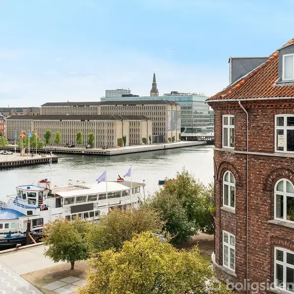 En hvid båd ligger til kaj ved en kanal. Til højre ses en ældre murstensbygning med buede vinduer. I baggrunden er moderne kontorbygninger og træer langs vandet.