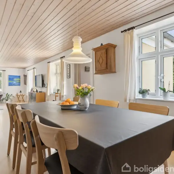 Et spisebord med seks stole er dækket med en dug og en vase med blomster. Handlingen er en spisestue i et lyst, moderne hjem med træloft og vinduer, der giver udsigt til haven.