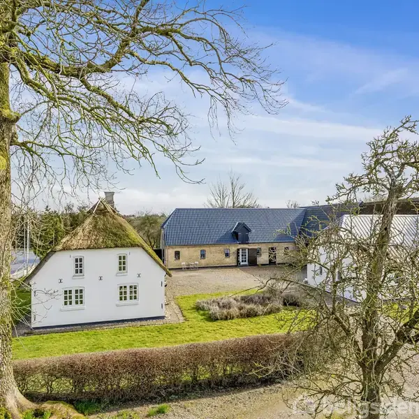 Et hvidt hus med stråtag står stille i et grønt landskab; omkring det er store træer og en anden bygning med sort tag.