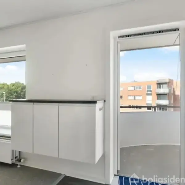 En hvid væg med et åbent vindue under et skab ses i et lyst rum. En åben dør fører til en altan med udsigt over bygninger og træer.