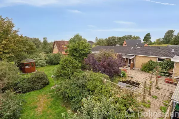 Et hus i et grønt område har en frodig have med buske og træer, en pergola og en græsplæne. Til venstre står en brun havepavillon.