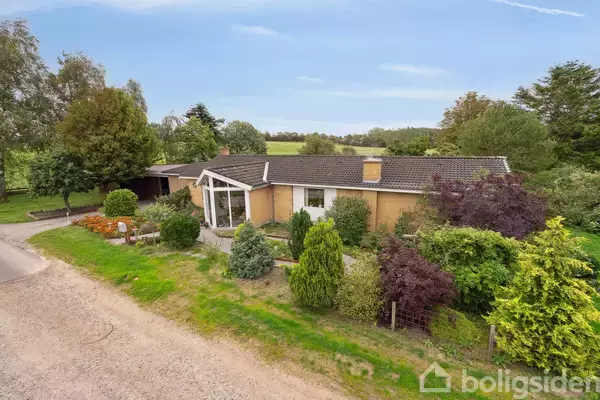 Et murstenshus med udestue står omgivet af frodige træer og buske i en landlig have. En grusvej løber langs venstre side af billedet.