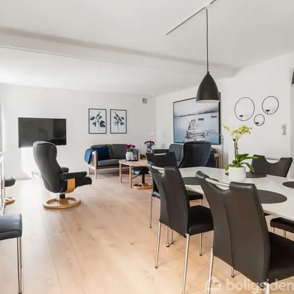 Spisestuesæt dominerer rummet; sorte stole står omkring et hvidt bord. Hyggelig kontekst med en stue i baggrunden, sofaer, vægkunst, tv og moderne belysning.