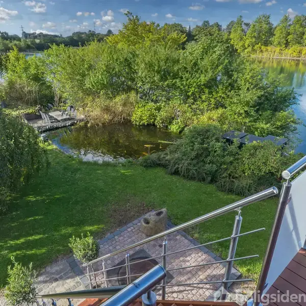 En balkon med metalgelænder giver udsyn over en velplejet have med terrasse, omkranset af træer. En flod løber langs kanten, omgivet af frodig vegetation og klar blå himmel.
