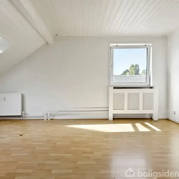 Et tomt rum med parketgulv. Lys strømmer ind gennem et tagvindue og et sidevindue. Væggene og loftet er hvide med en radiator under sidevinduet.