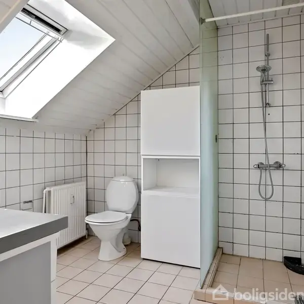 Toilet ved siden af bruseren i et badeværelse med skråloft. Fliser dækker væggene og gulvet, og en tagkvist tilføjer naturligt lys.