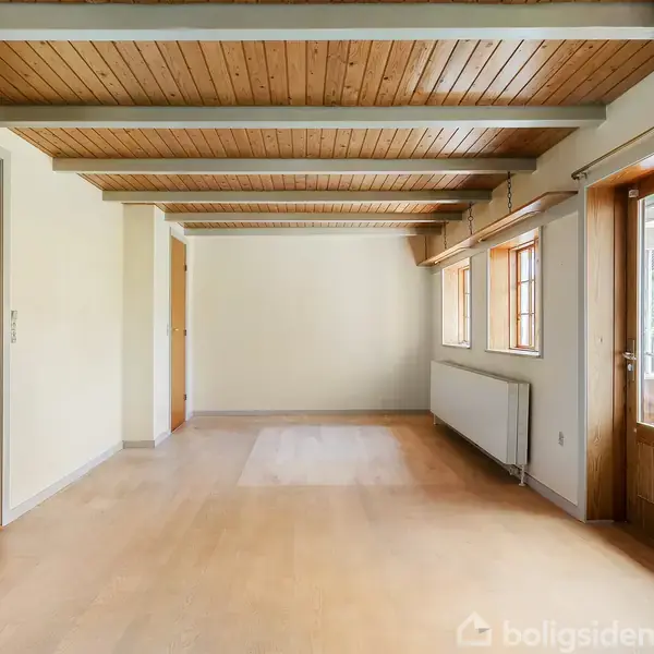 Træloft med synlige bjælker. Gulvet er lyst trægulv. Rummet har flere døre og vinduer, hvide vægge og åben forbindelse til en trappe. Radiator under vinduerne.