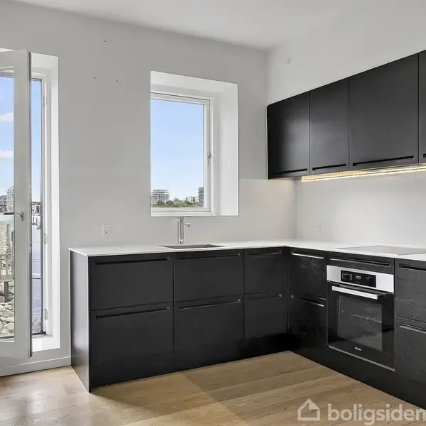 Et moderne køkken med sorte skabe og indbygget ovn oplyst af LED-belysning. Stort vindue og glasdør åbner ud mod en udsigt med bygninger og stensætning udenfor.