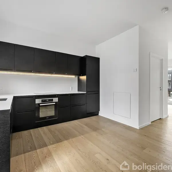 Sort køkken med indbyggede skabe og ovn indbygget i en lys, moderne lejlighed med trægulve, udført i minimalistisk stil. En åben dør fører til et rum med store vinduer.