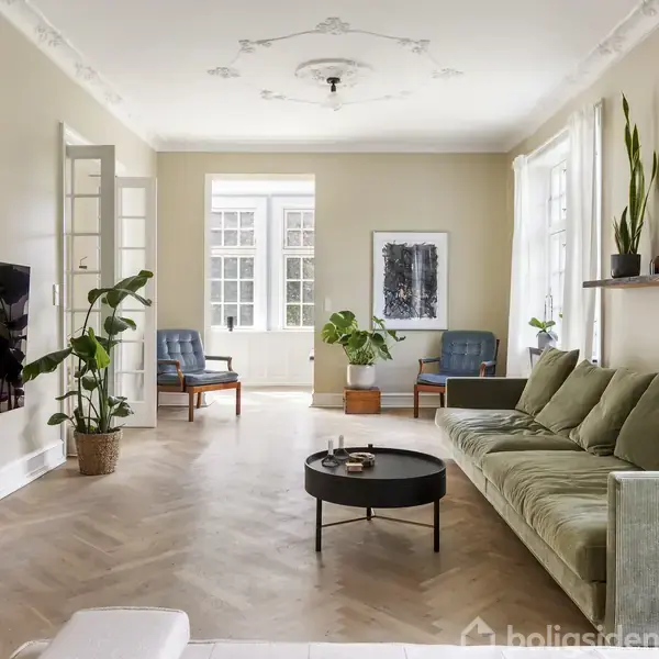 Stue med grøn sofa, flere planter og kunstværk på væggen. Et rundt sofabord står foran sofaen. Store vinduer fylder rummet med lys. To blå stole står i baggrunden.