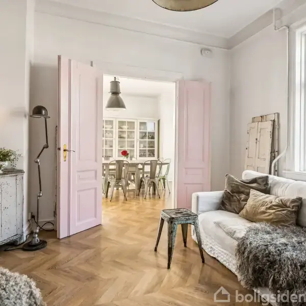 Stue med hvid sofa dækket af grå tæppe, lille bord foran. Til venstre er der et skab med lysestage. Åben dør mod spisestue med bord og stole. Trægulve.