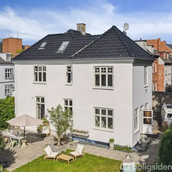 Hvidt to-etagers hus med sorte teglsten, omkranset af grønne buske. På terrassen står havemøbler, og der ses nabobygninger i baggrunden under en blå himmel.