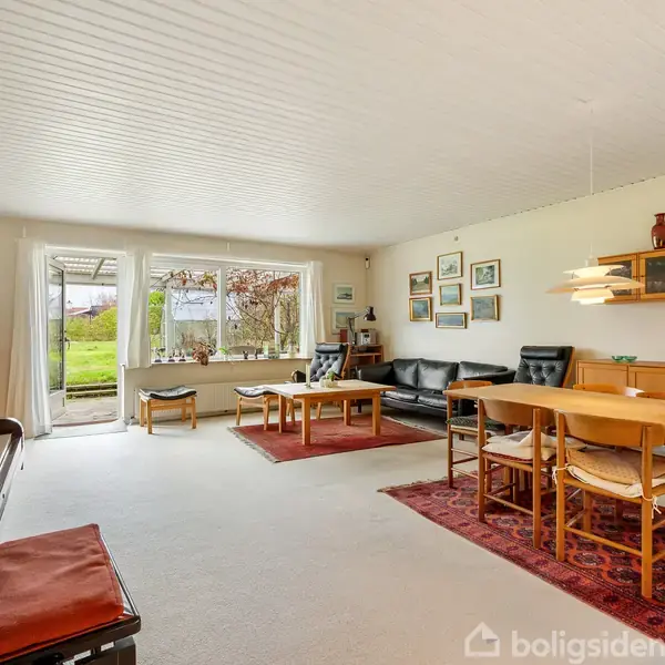 Et lyst, rummeligt stueområde med et klaver til venstre, sofaer og et sofabord i midten og et spisebord til højre. Stort vindue giver udsigt til en grøn have.