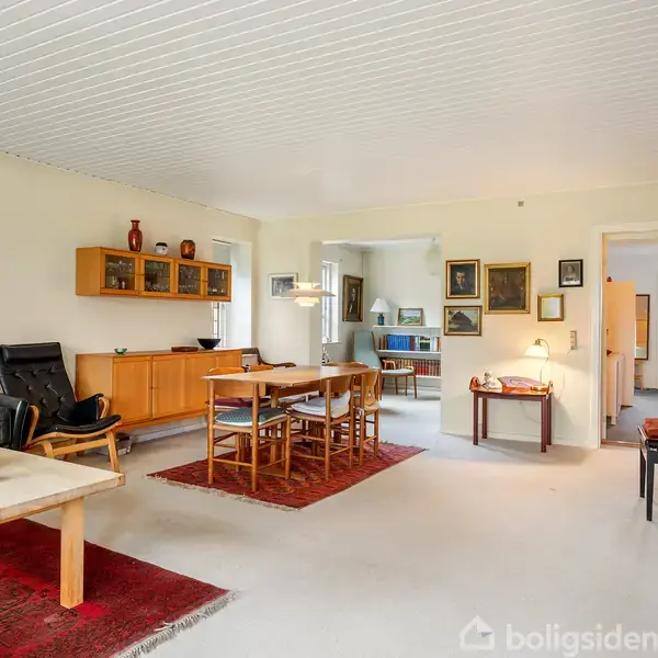 Stue med sort lænestol og spisebord, omgivet af lyse vægge med kunstmalerier. På væggen hænger et skab. Der er også et klaver og flere tæpper på gulvet.