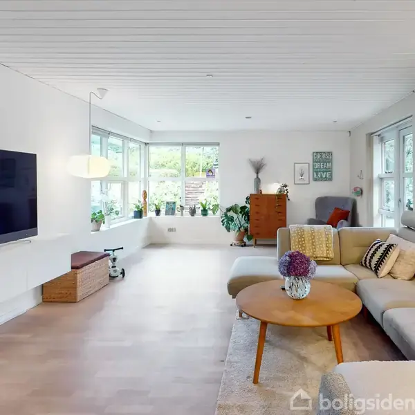 Stue med lys sofa og runde træbord, stor vægmonteret TV, planter og dekorationer i lys, moderne indretning. Store vinduer i baggrunden giver naturligt lys og udsigt til haven.